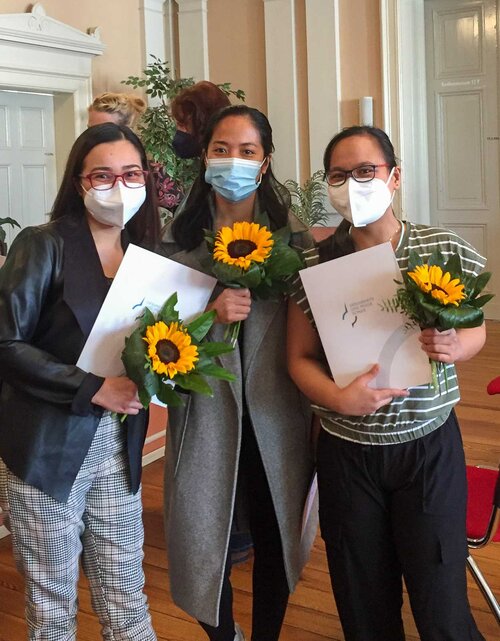 Drei Frauen stehen mit Zeugnispapieren und Sonnenblumen zusammen und schauen erfreut 