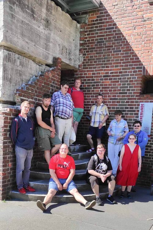 Eine Gruppe Menschen hat sich auf Stufen gesetzt vor einem alten, halb verfallenen großen Industriegebäude aus Backstein